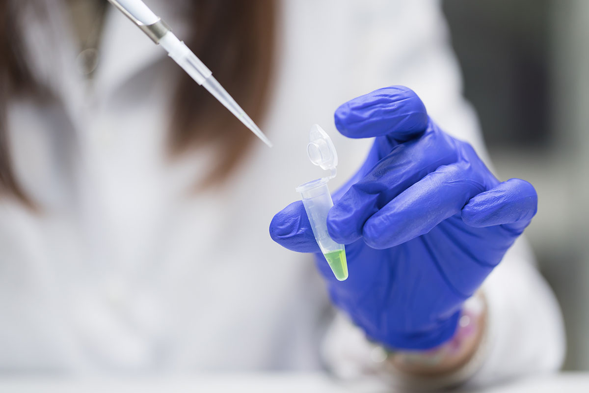 A gloved hand holding an open microcentrifuge tube of green liquid beneath a pipette tip in a research laboratory