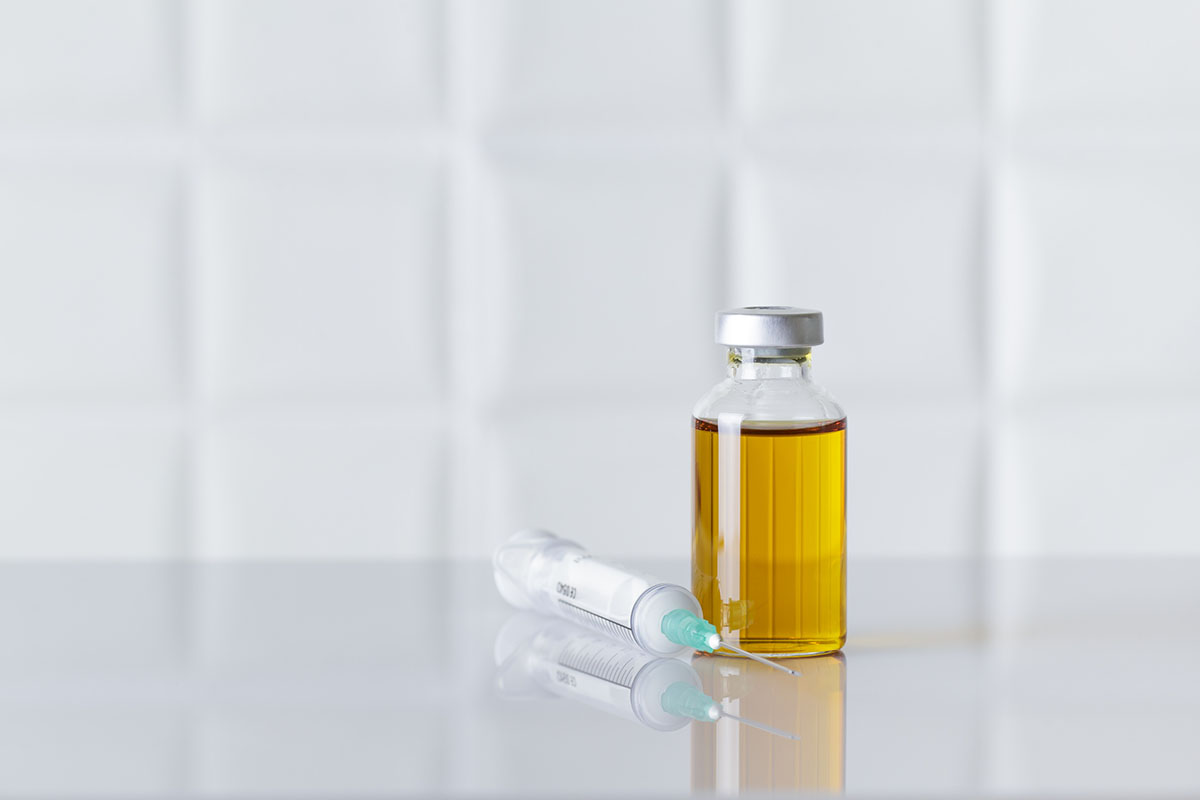 A glass vial of amber injectable medicine beside a syringe with a teal needle cap on a white laboratory surface